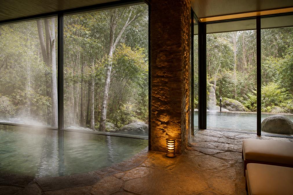 Serene luxury spa sanctuary with natural thermal pools, warm stone surfaces, soft ambient lighting, and floor-to-ceiling windows overlooking pristine Tasmanian bushland
