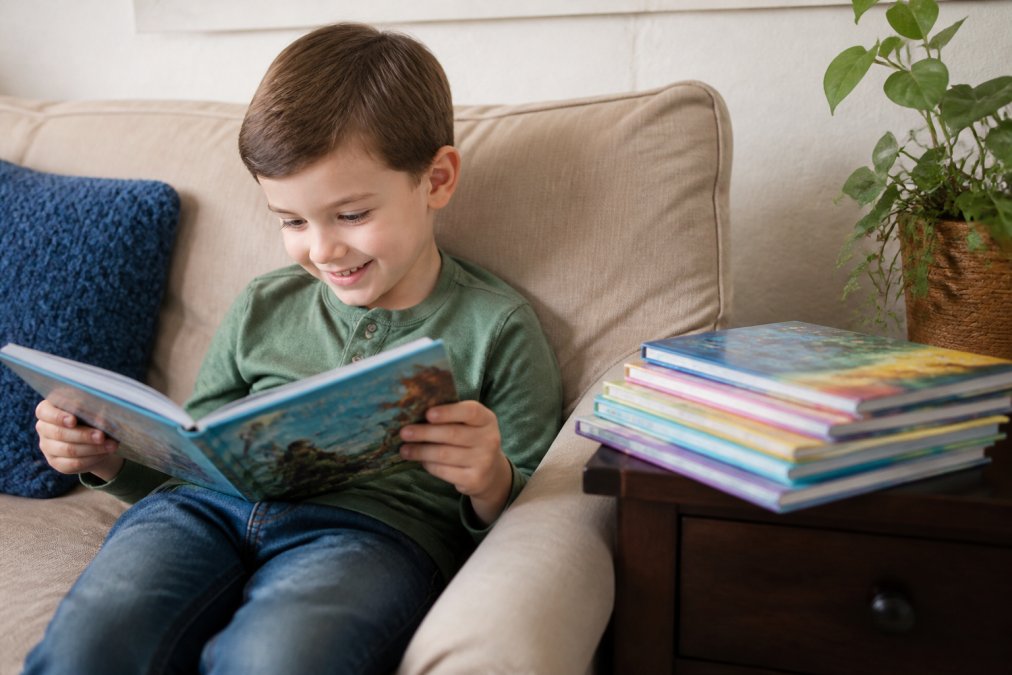 A child sitting on a couch at home, holding an open picture book and smiling while reading, with several colorful adventure books stacked on the side table