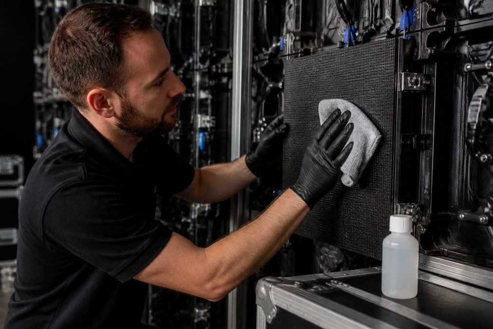 Technicus met beschermingshandschoenen voorzichtig een LED-module reinigend met microfiber doek, reinigingsmiddel in klein flesje naast zich, LED-scherm zachtjes verlicht op achtergrond