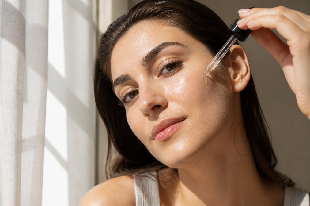 Close-up of a woman's face with radiant, even-toned skin, applying a serum with a dropper, soft natural light, clean background
