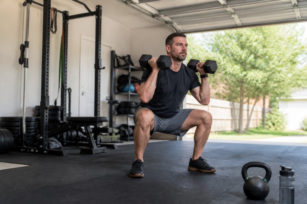 A fit father in his late 30s performing a dumbbell squat in a bright, simple home garage gym while a timer on the floor shows a countdown