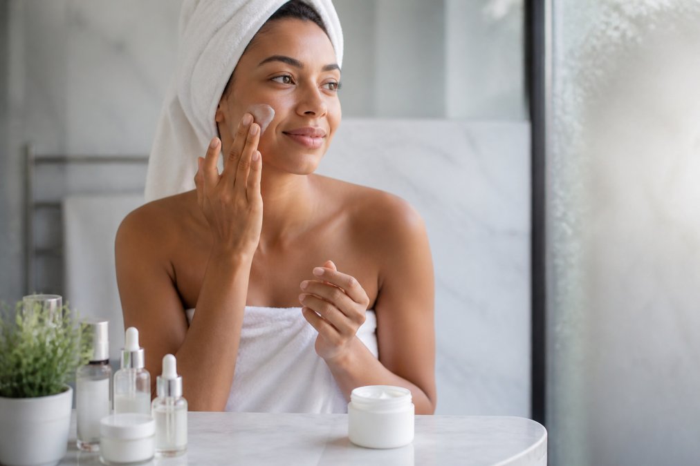 A woman gently applying a lightweight moisturiser to her face in a bright, minimal bathroom, surrounded by a few clean skincare products arranged neatly on a white marble shelf, soft natural morning light coming through a frosted window