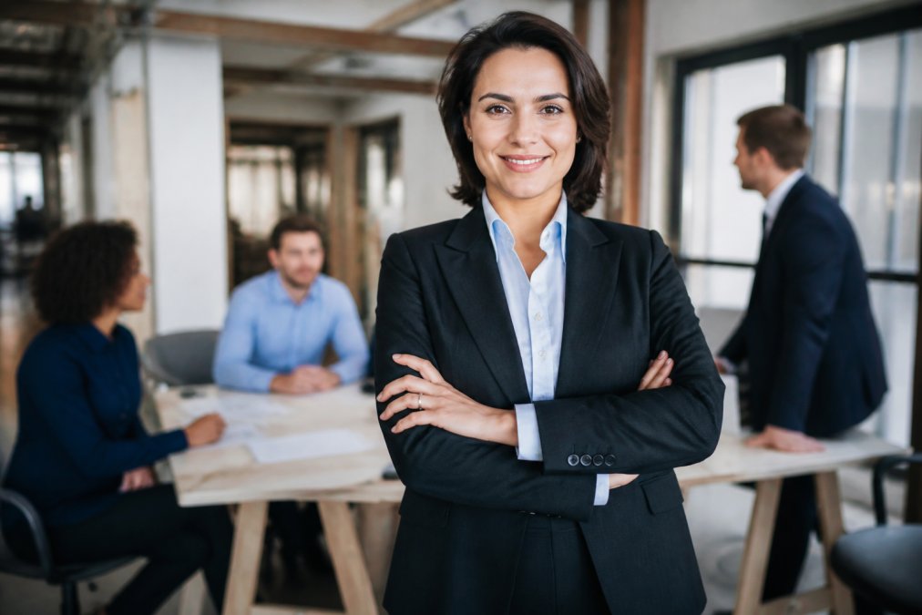 Professional woman in business attire standing confidently with shoulders back, making direct eye contact, in a modern workplace setting with colleagues visible in the background