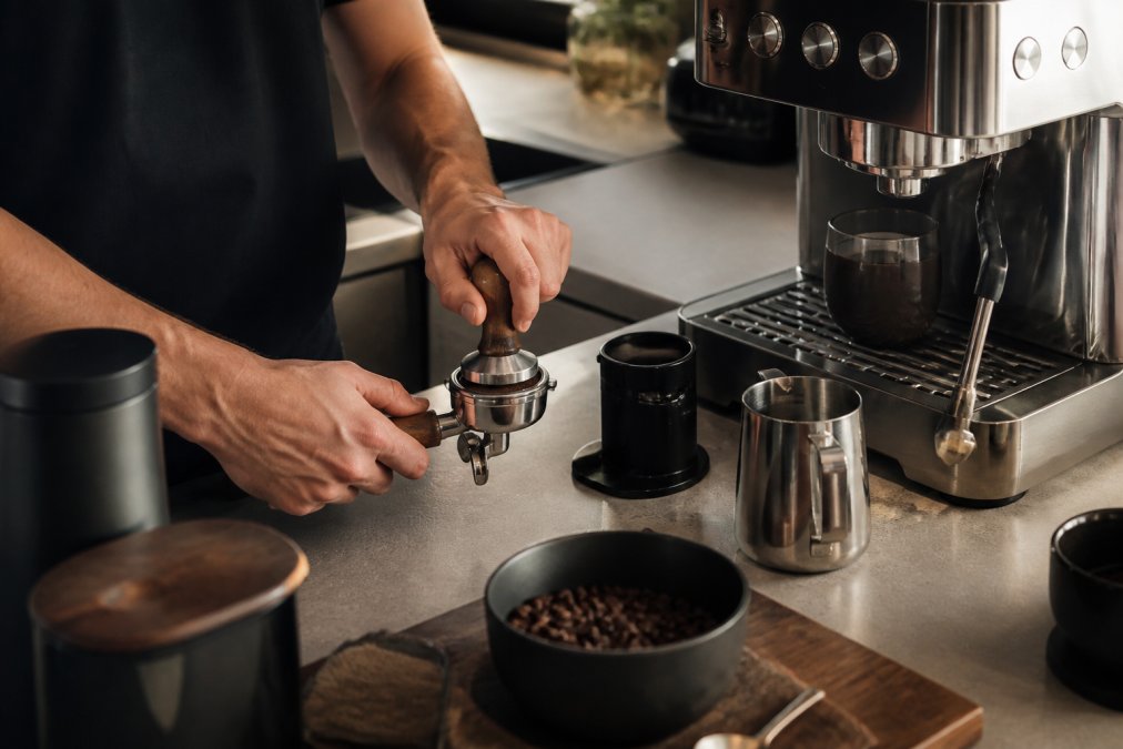 Nærbillede af en hjemmebarista, der bruger en tamper og mælkeskummer ved en espressomaskine, med gourmetkaffe og stilfuldt udstyr på køkkenbænken