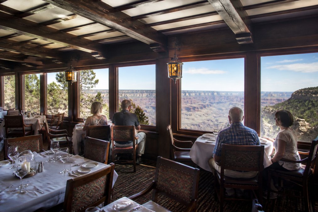 Sunlit dining room inside a historic national park lodge, dark wood beams, white tablecloths, large windows framing a canyon vista, a few diners seated near the windows