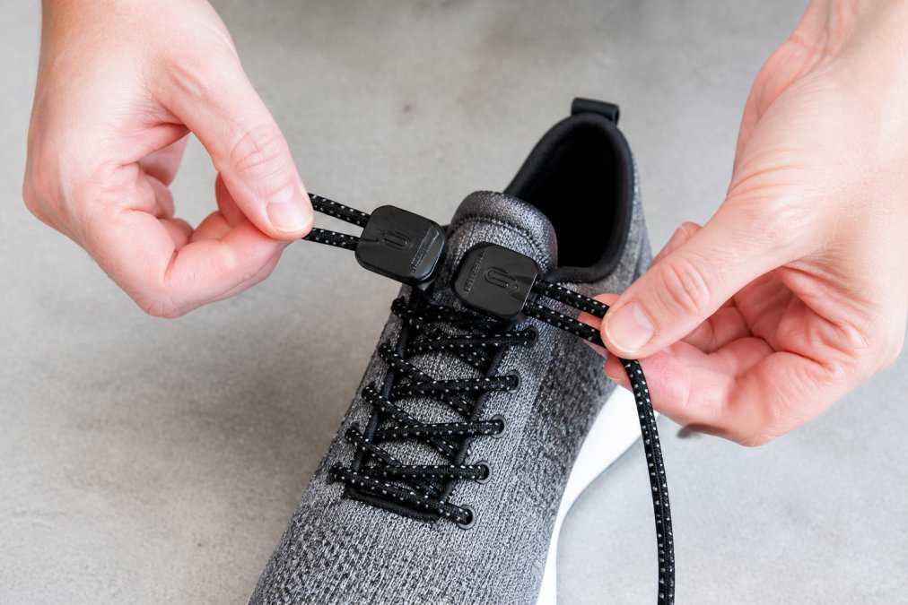 Close-up of hands threading elastic laces through shoe eyelets during installation, showing the push-and-pull method with a sport shoe on a flat surface, natural lighting from above