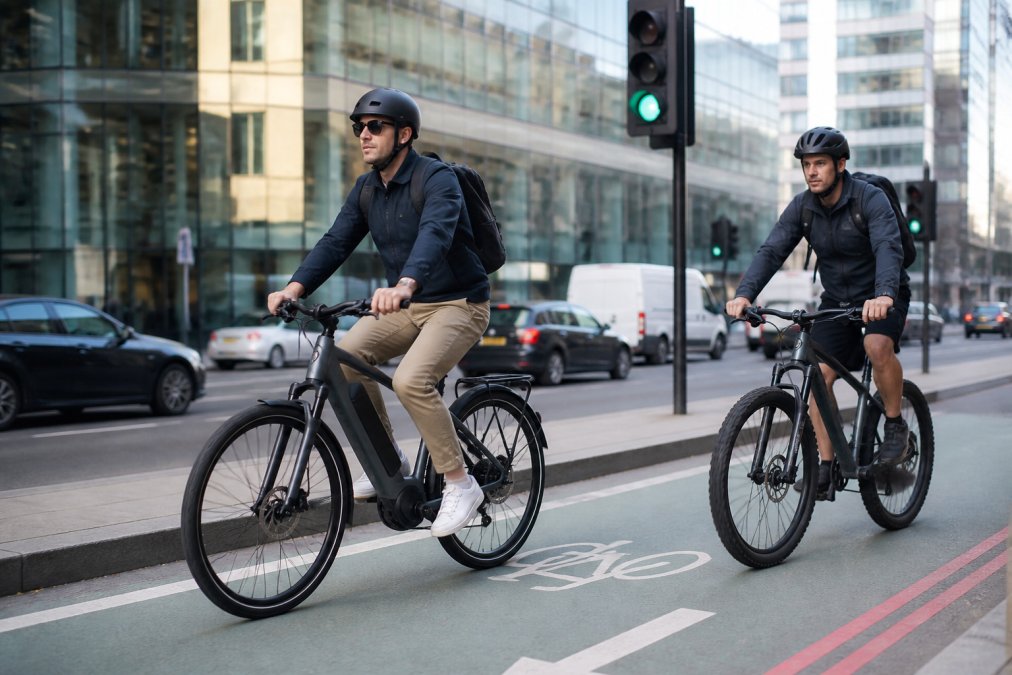 Urban commuter riding an electric bike on a city street with dedicated bike lanes, traffic lights, and office buildings in the background, shot during morning rush hour with natural daylight