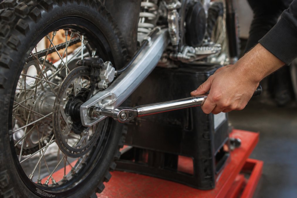 Close-up of a mechanic's hands using a torque wrench on the axle bolt of an electric dirt bike in a workshop, with the bike elevated on a stand