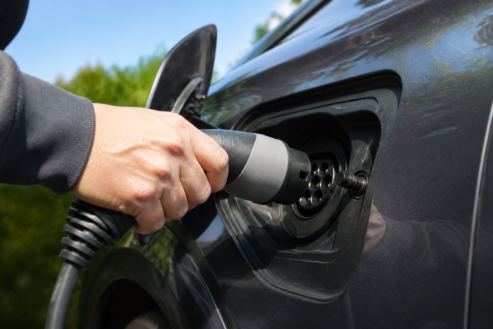 Close-up of hands holding a Type 2 charging connector being inserted into an electric vehicle's charging port on a sunny day, showing the precise connection point