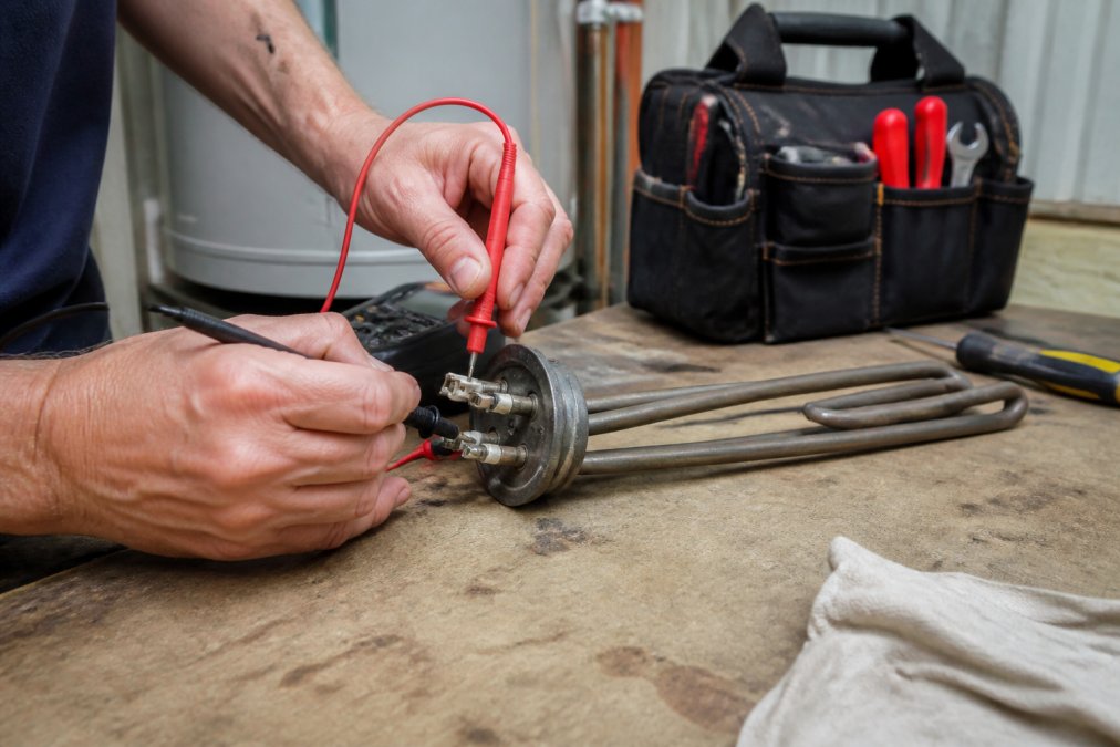 A close-up of a homeowner's hands using a digital multimeter to test the terminals of an electric water heater heating element that has been removed from the tank, with the element resting on a workbench