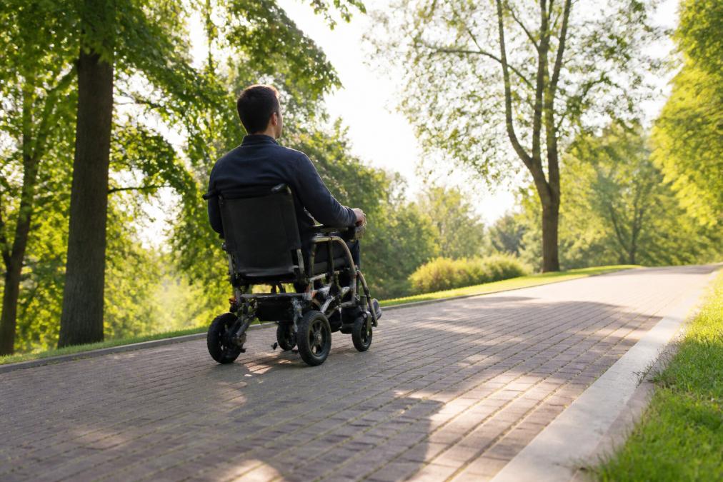 Person in lightweight folding electric wheelchair navigating a paved park path with a gentle incline, trees and natural sunlight in background, demonstrating real-world terrain conditions
