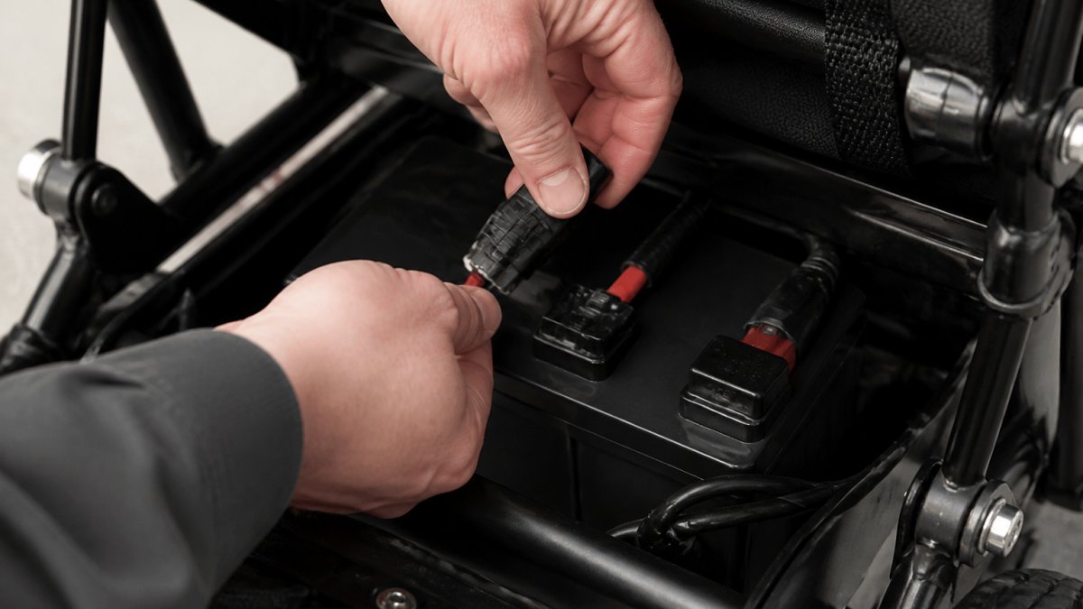 Close-up of hands carefully disconnecting a battery from the power connection ports on an electric wheelchair battery compartment, showing the F1 and F2 terminals clearly