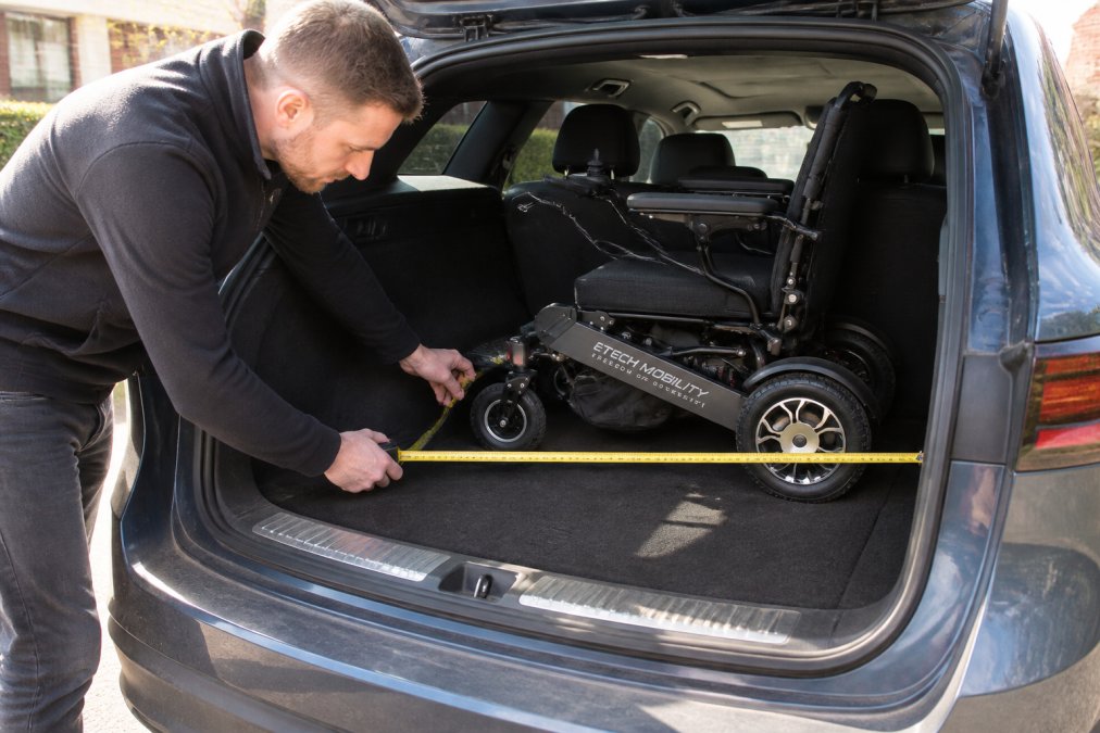 Person measuring the interior of a car boot with a rigid tape measure, carefully noting the depth from opening to back wall and the width between wheel arches, with the boot lid fully open in daylight