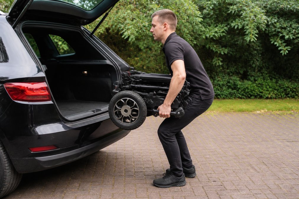 Person demonstrating proper lifting posture while placing a folded electric wheelchair into a car boot, keeping the chair close to the body with bent knees and neutral spine, viewed from the side with natural outdoor lighting
