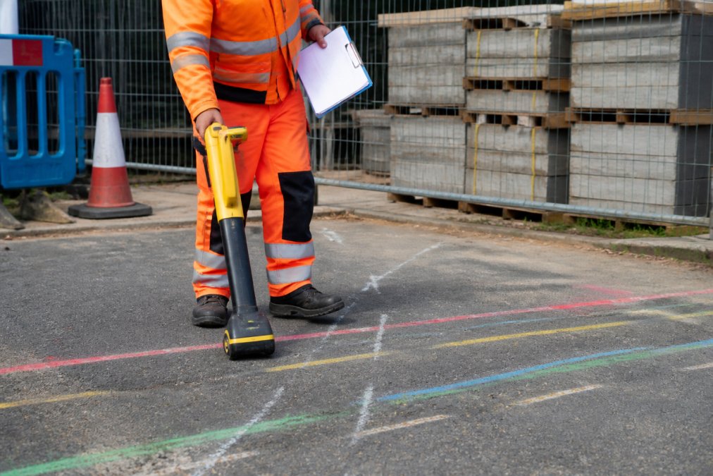 Groundworker using a Cable Avoidance Tool (CAT) to scan marked ground near coloured service route markings, with a permit-to-work document visible on a clipboard in bright daylight
