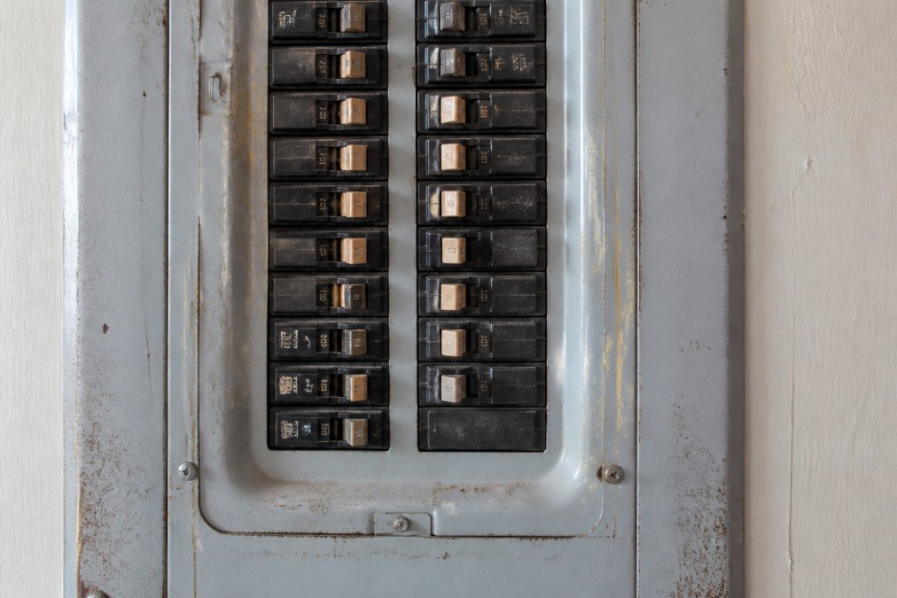 Close-up view of an electrical panel box with visible circuit breakers, showing wear on the metal housing and older-style breaker switches in an installed residential setting