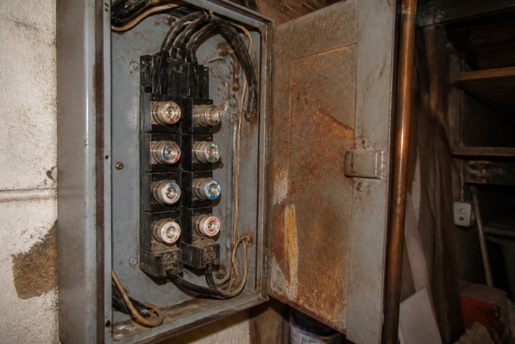 Close-up of an aged residential electrical panel with outdated glass fuses, discolored wiring, and visible corrosion inside the panel door, illuminated by a work light in a dim utility room