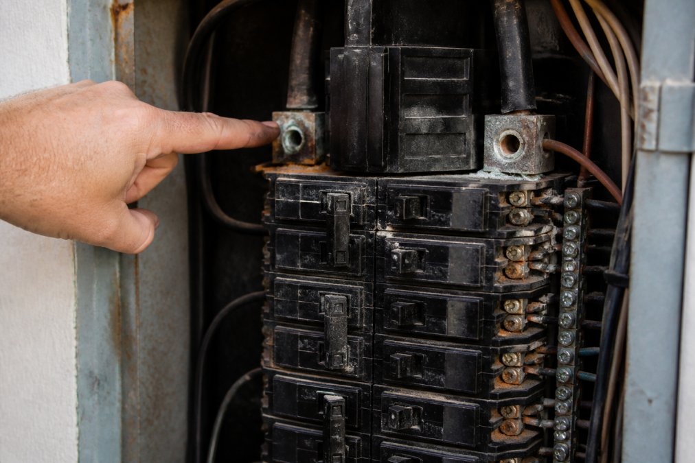 Close-up of an electrical panel with multiple circuit breakers showing visible corrosion and wear, with a licensed electrician's hand pointing to a corroded main breaker connection