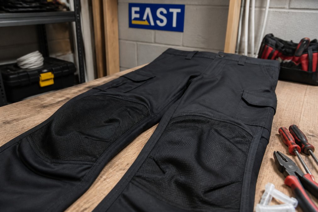 Close-up of a pair of work trousers showing reinforced knee-pad pockets and durable stitching, laid on a workbench with tools nearby