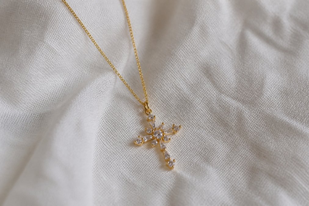Close-up of delicate cross necklace with fine chain draped on soft white linen fabric, showing intricate pendant details and polished gold finish in natural daylight