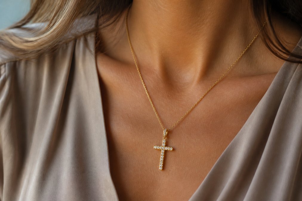 Close-up of a gold pavé cross pendant resting on a woman's collarbone over a neutral-toned blouse, soft natural lighting highlighting the sparkling CZ stones