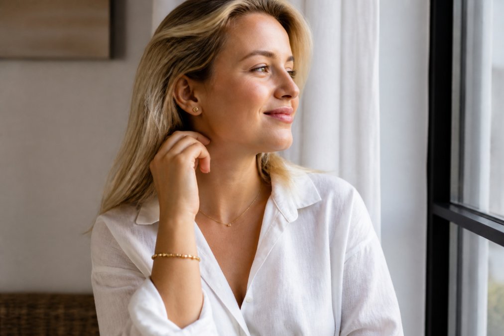 Vrouw met blonde haar in een casual witte linnen blouse draagt gouden oorknopjes, een dunne gouden ketting en een gouden armband, gefotografeerd in natuurlijk daglicht bij een raam