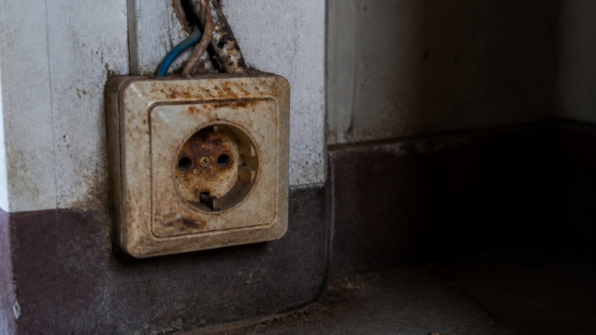 Close-up van een oud stopcontact met zichtbare roest, verkleuring en losse draden in een donkere hoek van een oudere woning