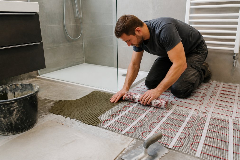 Vakman die een dunne verwarmingsmat voorzichtig onder tegels in een moderne badkamer installeert, met tegellijm en gereedschap zichtbaar op het werkoppervlak