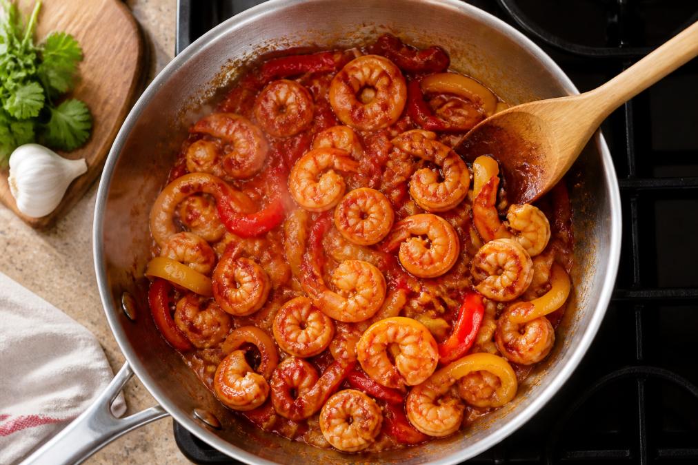 Overhead shot of a colourful one-pan dinner with golden-brown shrimp, vibrant red and yellow peppers, and glossy spicy sauce simmering in a stainless steel skillet, steam rising gently, wooden spoon resting on the rim, warm kitchen lighting