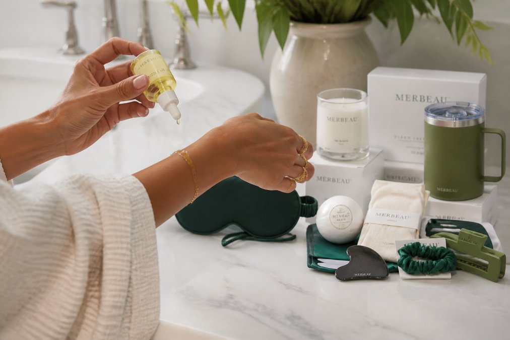 Close-up of hands applying luxurious natural body oil to skin, with wellness products like bath salts, a scented candle, and a soft towel arranged on a clean marble surface nearby