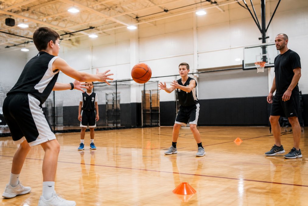 Young basketball players in a gymnasium practicing fundamental drills under coach supervision, executing passing and footwork exercises with focused intensity under bright overhead lighting