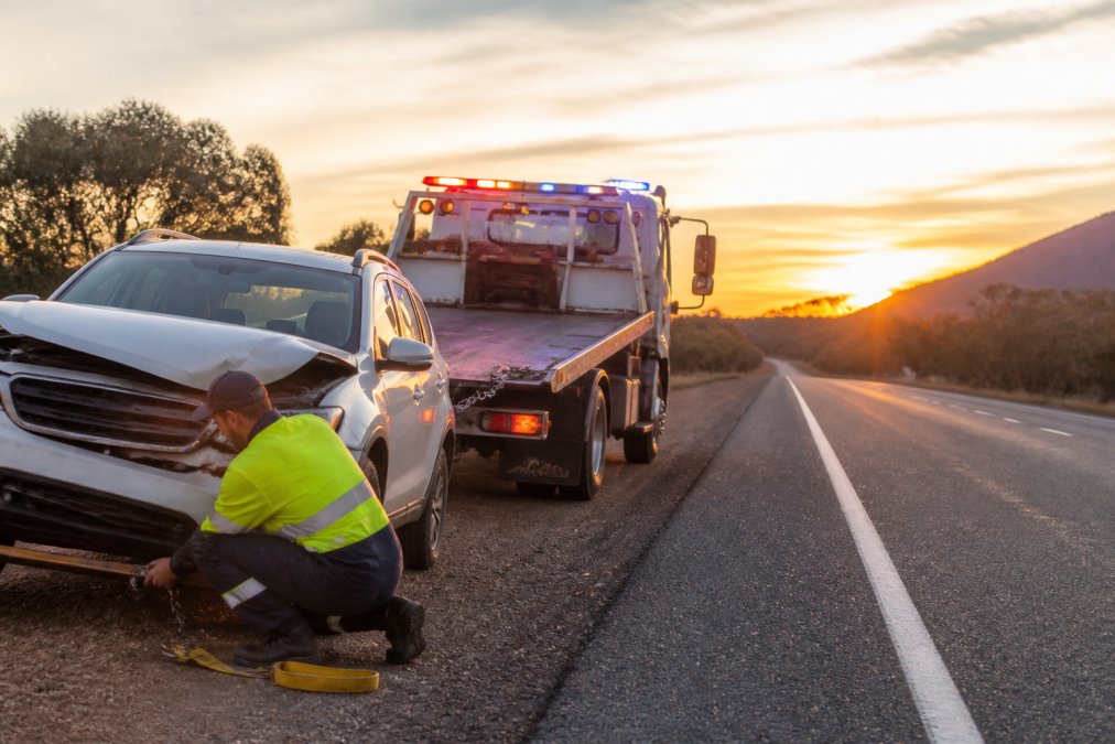 Tow truck arriving at damaged vehicle on regional road with emergency lights visible and operator preparing recovery equipment, late afternoon lighting