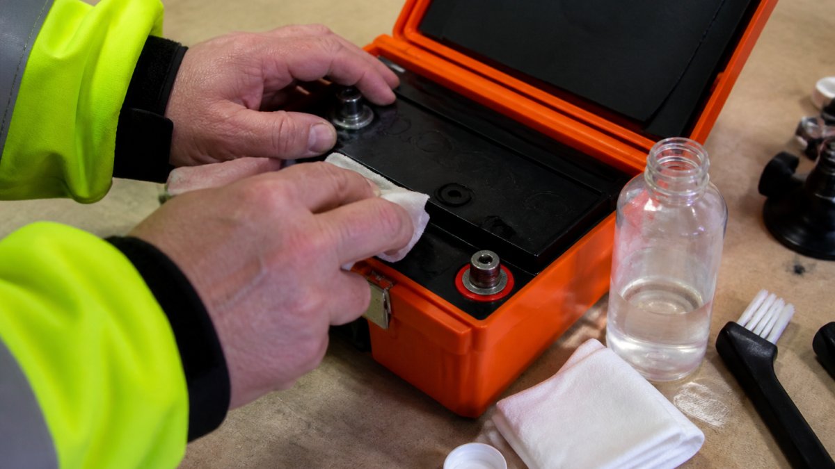 Emergency Beacon Battery Maintenance for Winter Close-up of hands carefully inspecting emergency beacon battery terminals and connections with a soft cloth and distilled water nearby on a clean workbench, natural workshop lighting