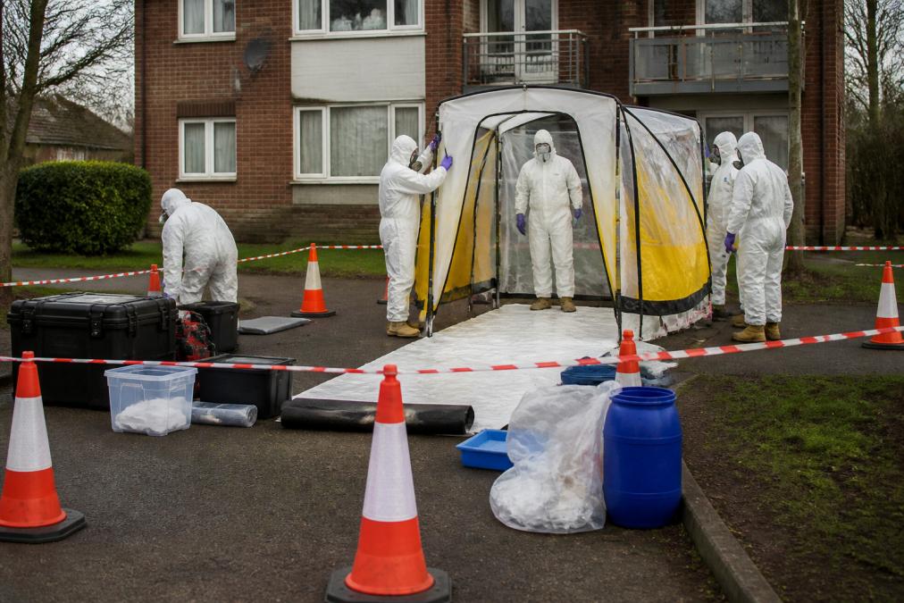 Fully suited decontamination technicians in white protective clothing and respirators setting up a decontamination shelter outside a residential housing block in overcast daylight, with safety cones and hazard tape marking the exclusion zone