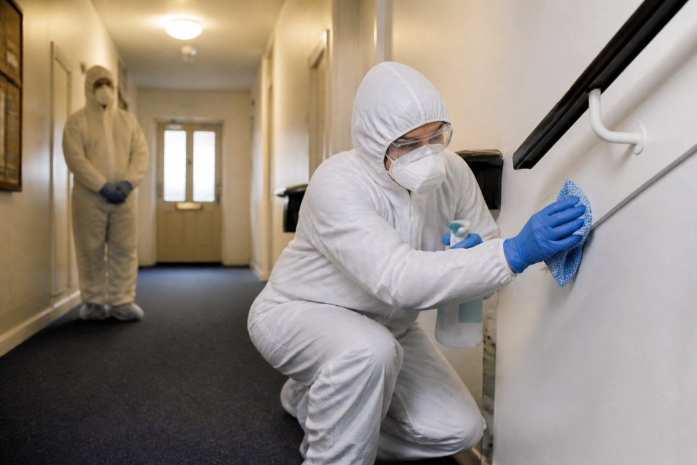 Staff member in full PPE performing decontamination training in a residential hallway while a colleague observes.