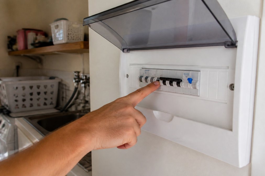 Close-up of a person's hand pointing to a tripped safety switch on a white domestic switchboard panel in a well-lit laundry room
