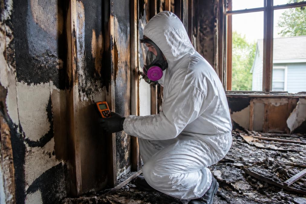 Professional restoration technician in full protective gear assessing fire-damaged residential walls with visible char and debris, using moisture meter in natural daylight from damaged window