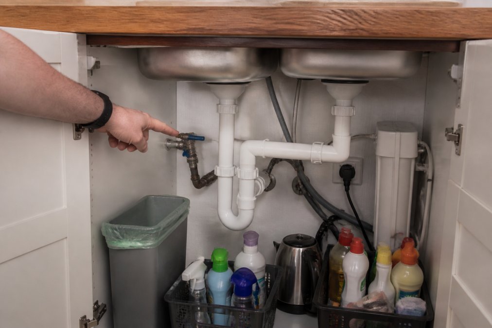 Homeowner's hand pointing to a stopcock isolation valve clearly visible in the cabinet beneath a kitchen sink, with pipes visible in the background