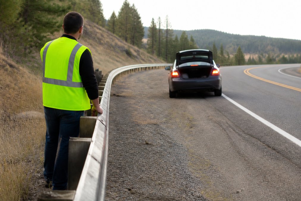 A driver in a reflective safety vest standing behind the guardrail of a rural highway, away from a parked sedan with its hazard lights on, waiting for assistance