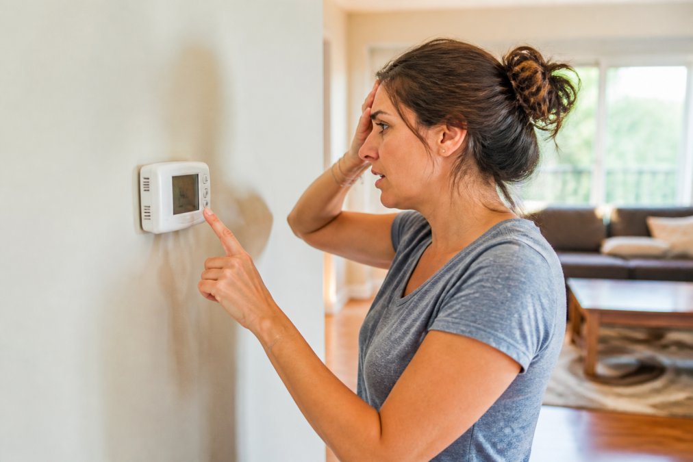 A homeowner standing in front of a wall-mounted thermostat with a concerned expression, checking the temperature display in a warm, sunlit living room with visible discomfort