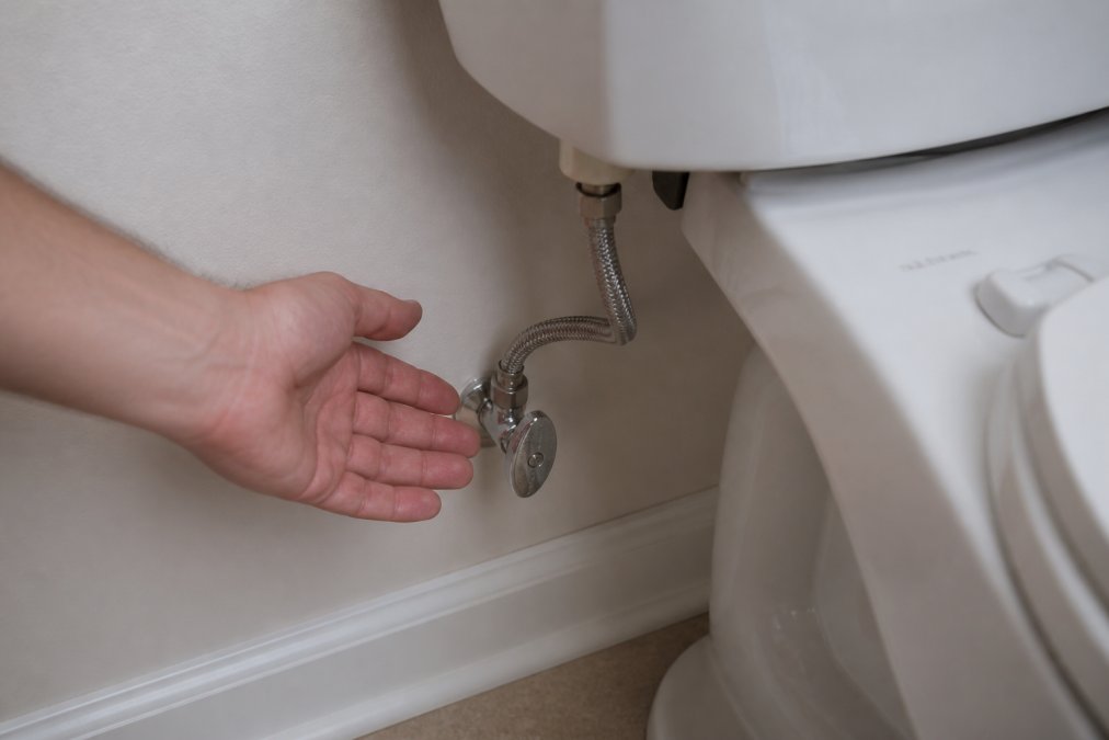 Close-up of a hand reaching toward the toilet tank, showing the shut-off valve location on the water supply line behind the toilet, with clear view of the valve handle and connection point