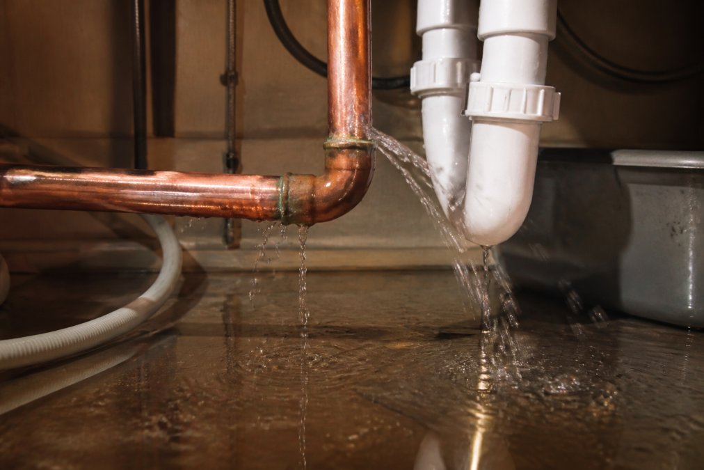 Close-up of water spraying from a burst copper pipe under a kitchen sink, water pooling across the cabinet floor in a dimly lit under-sink space