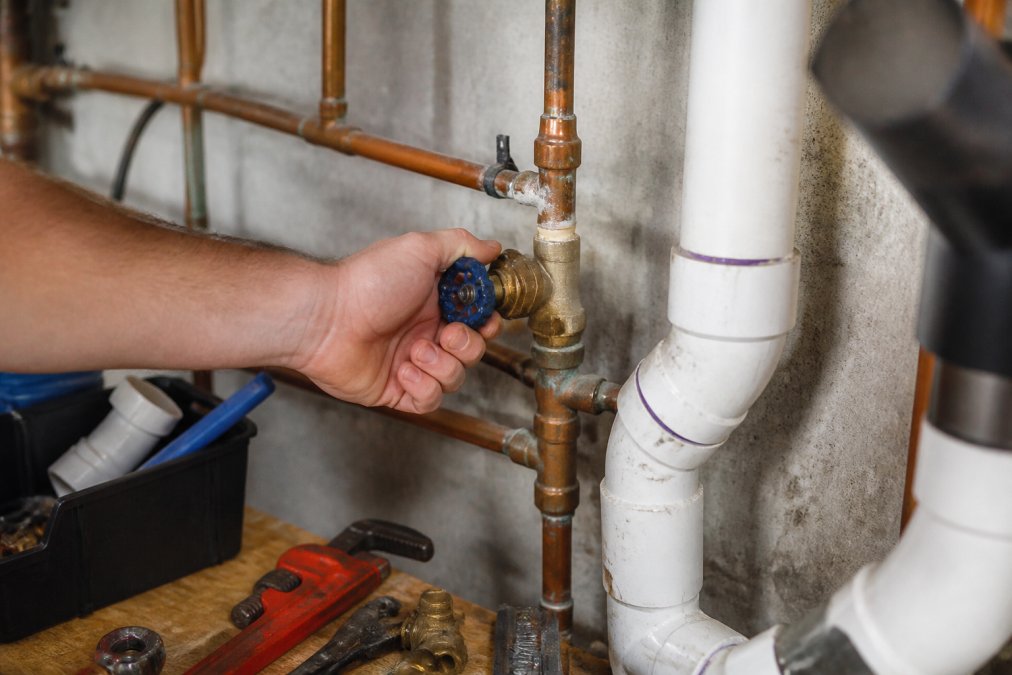 Homeowner's hand gripping the main water shut-off valve handle in a basement utility area, with copper and PVC pipes visible in the background, natural basement lighting