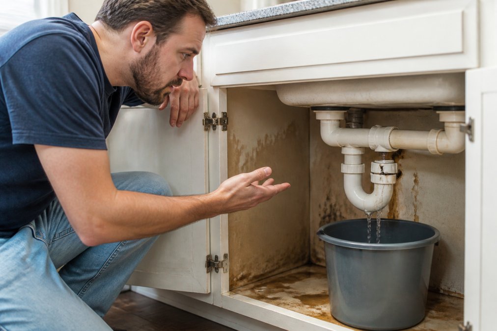 Homeowner in casual clothing kneeling under a kitchen sink, examining a visible burst pipe with water actively dripping into a bucket, water damage visible on cabinet interior, concerned expression on face