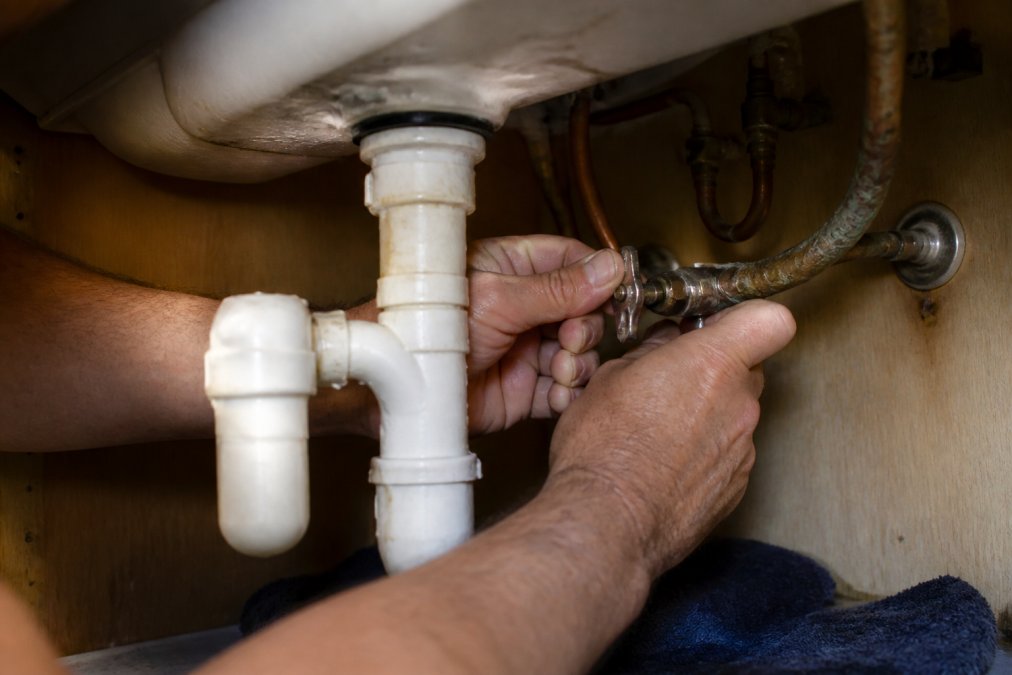 Close-up of a homeowner's hands turning off a residential water shut-off valve under a bathroom sink, with water-stained pipes and copper fittings visible in the background under dim under-sink lighting