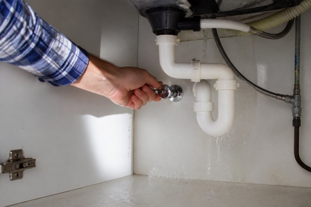 Homeowner turning off the main water shut-off valve under the sink, with water actively dripping from a burst pipe visible in the background