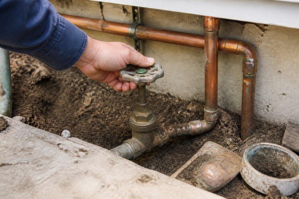Homeowner's hand turning a water main shut-off valve located near the foundation of a house, with copper pipes visible in the background and soil around the valve