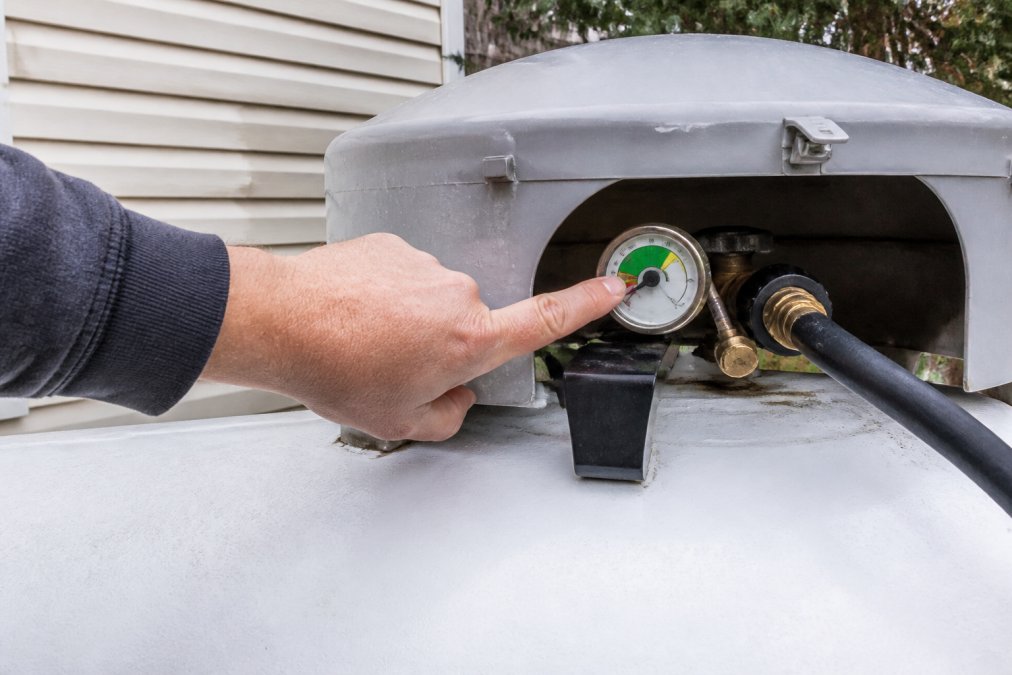A homeowner's hand pointing to the propane tank gauge dial on an outdoor residential tank, showing the float indicator in the green zone with clear visibility of the percentage markings in natural daylight