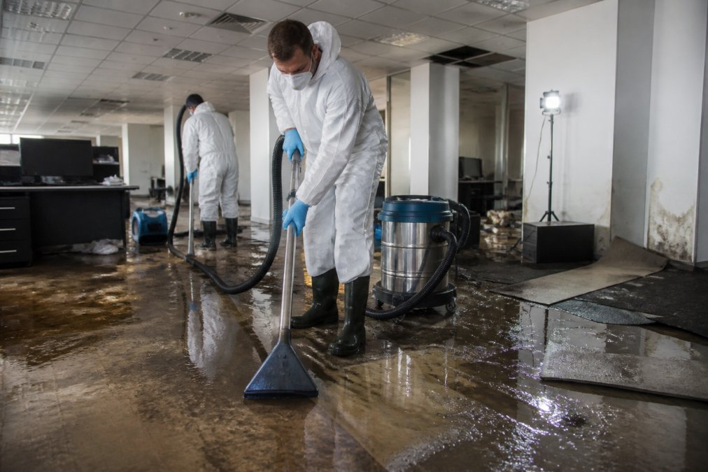 Restoration technicians in white protective suits using industrial water extraction equipment inside a flooded commercial office, with damaged flooring, displaced furniture, and emergency lighting