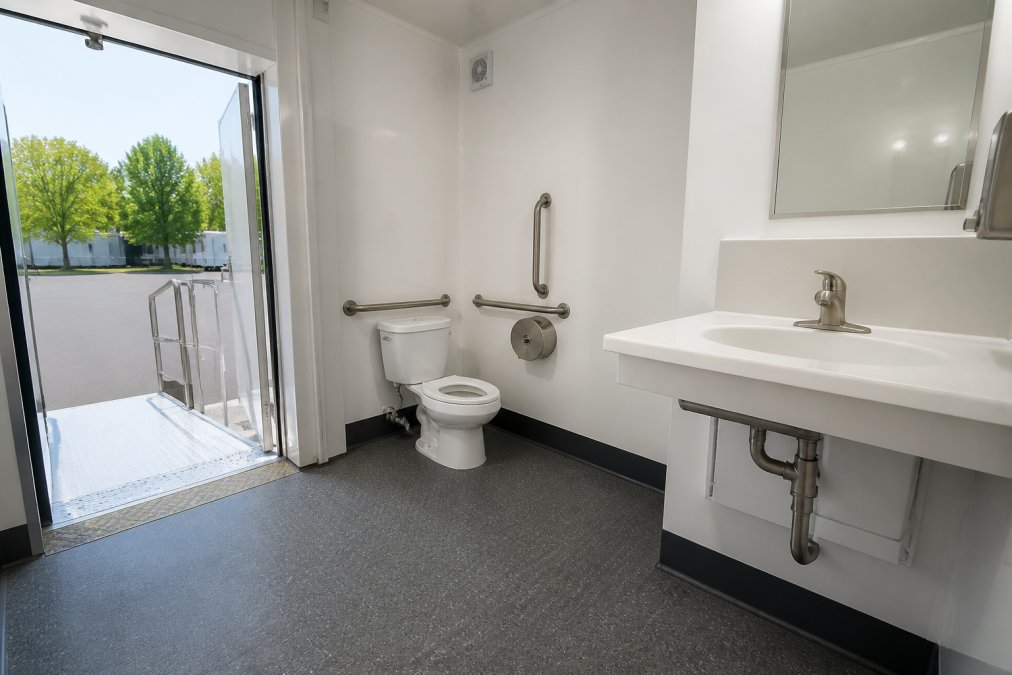Interior of an ADA-accessible restroom trailer showing stainless steel grab bars on both sides of a porcelain toilet, a wide open doorway with a low-threshold entry, a clean white sink with lever-style faucet, and bright overhead lighting with slip-resistant flooring